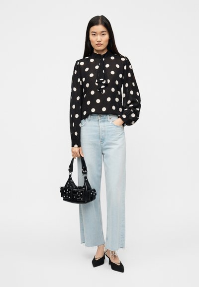 Μαύρη μπλούζα με μακριά μανίκια και λευκά πουά, με λεπτομέρεια βολάν, σε συνδυασμό με ανοιχτό μπλε παντελόνι φαρδύ, μαύρη τσάντα και μυτερά μαύρα παπούτσια.