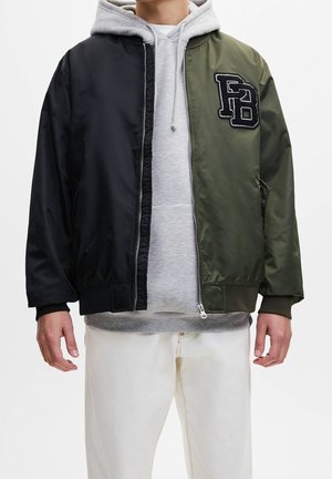 Homme portant une veste bomber noire et vert olive par-dessus un sweat à capuche gris et un pantalon blanc, debout devant un fond blanc uni.