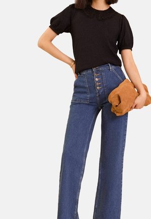 Femme portant un chemisier noir à manches courtes avec un col en dentelle, un jean bleu taille haute avec des boutons, tenant une pochette marron.