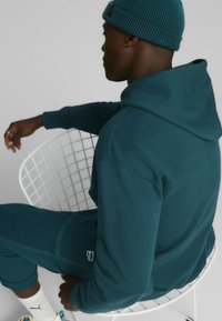 Sudadera de algodón color azul verdoso con cordón y puños acanalados, combinada con pantalones de chándal a juego, sentado en una silla de alambre blanca. Lleva un gorro en la cabeza.