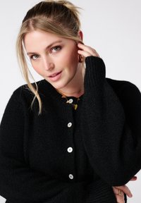 Zwarte gebreide cardigan met een gestructureerde stof, een ronde halslijn en witte knopen. Het ontwerp heeft een subtiele oversized pasvorm en geribbelde manchetten.