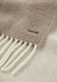 Beige wollen sjaal met franjes, met een soepele textuur en een decoratieve pin in zilverkleur met een oranje accent.