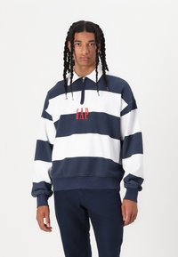 Sudadera a rayas en azul marino y blanco con cuello con cremallera, presenta un logo rojo "GAP" en el pecho. Material de algodón, corte relajado.