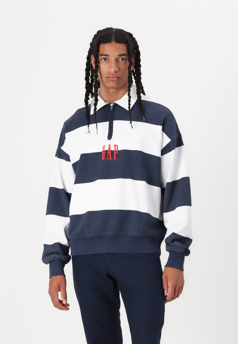 Sudadera a rayas en azul marino y blanco con cuello con cremallera, presenta un logo rojo "GAP" en el pecho. Material de algodón, corte relajado.