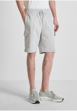 Mann trägt hellgraue Cargo-Shorts mit Kordelzug in der Taille und hellgraue Sneaker, steht mit Händen in den Taschen.