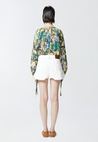 Cvećasta bluzica s dugim rukavima, višebojnim tropskim uzorkom, u kombinaciji s bijelim šortsem i bež natikačama. Pogled straga.