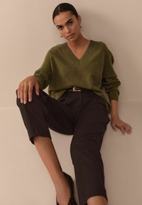 Olive green v-neck sweater, loose fit, paired with high-waisted black trousers. Bold, textured black shoes with a leopard print accent.