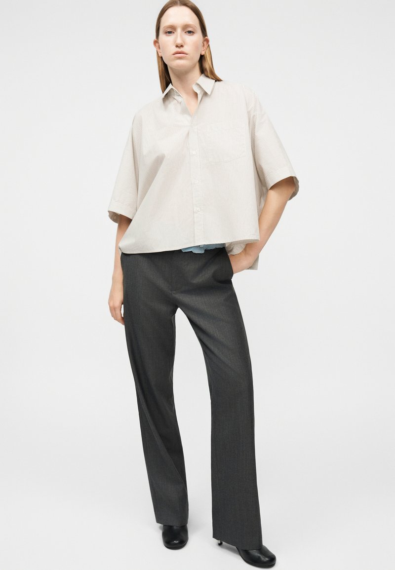 Jonge vrouw met een losse beige korte mouwen blouse met knopen, donkergrijze rechte broek en zwarte schoenen met hak, staand tegen een effen achtergrond.