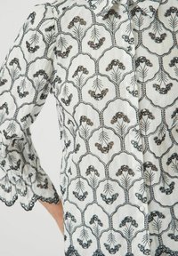 Witte stoffen shirt met zwarte en oranje bloemenborduursels, voorzien van een knoopsluiting aan de voorkant en een gegolfde zoomdetail op de mouwen.