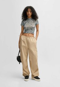Taillierte, verkürzte Bluse mit einem Druck in Schwarz, Grün und Weiß; kombiniert mit lockeren beigen Hosen und schwarzen Sneakers mit weißen Sohlen.