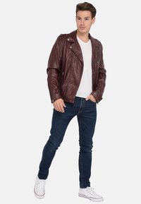 Veste de moto en cuir bourgogne avec fermeture éclair asymétrique, col à pression et poches latérales, associée à un jean en denim bleu et des baskets blanches.