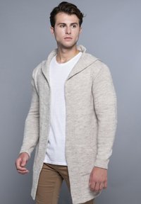 Duga, svijetlosmeđa pletena kardigan jacket s otvorenim prednjim dijelom, velikom kapuljačom i rebrastim manžetama. U kombinaciji s bijelom majicom i kašmir hlačama.