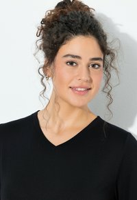 Top negro con cuello en V y textura suave y lisa. El modelo tiene el cabello rizado y lleva pendientes pequeños de aro. Vista frontal con fondo claro.