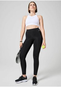 Valkoinen urheilu crop top korkealla kauluksella, yhdistettynä korkeavyötäröisiin mustiin legginsseihin. Malli pitää kädessään tennismailaa ja palloa. Mustat lenkkarit.