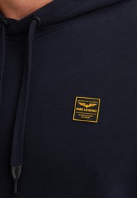 Sudadera azul marino hecha de tela suave, con capucha ajustable y un parche cuadrado de logo amarillo y negro en la parte frontal.