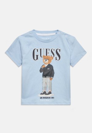 Camiseta azul claro para niño pequeño con un osito de peluche que lleva una chaqueta negra y pantalones a rayas, texto "GUESS" arriba y "Los Angeles Est. 1981" abajo.