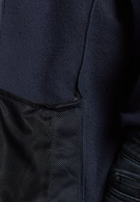 Marineblaues Fleece-Jackett mit einer schwarzen Mesh-Tasche, strukturiertem Stoff und fertiggestellten Nähten, entworfen für Komfort und Atmungsaktivität.