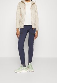 Vaaleanbeige pehmeä takki hupulla, yhdistettynä tummansinisiin legginsseihin, joissa on mustat yksityiskohdat, ja vaaleanvihreisiin urheilukenkään.