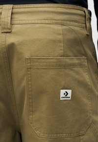 Κοντινό πλάνο πίσω τσέπης χακί παντελονιού με λευκή ετικέτα λογότυπου Converse ραμμένη στην τσέπη.