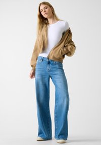 Bež bomber jakna na patent, bijela uska majica i svijetloplave široke traperice. Glatki materijal, ležeran dizajn i opušteni kroj.