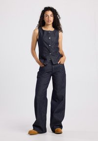 Navy sinistä raidoitettua denimiä oleva asu, johon kuuluu hihaton napitettava paita ja leveälahkeiset housut, joissa on tekstuuri ja minimalistinen muotoilu.