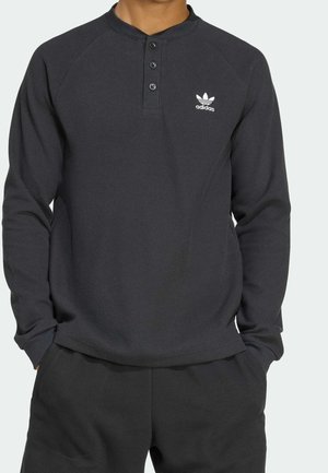Mand iført mørkegrå, tekstureret henley-langærmet skjorte med to knapper og hvidt Adidas-logo på brystet.