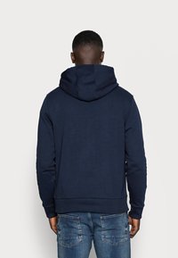 Man som bär en marinblå hooded sweatshirt och blå jeans står vänd bort från kameran mot en ljus, enfärgad bakgrund.