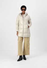 Lichte, crèmekleurige pufferjas met horizontale stiksels, ritsluiting en een capuchon. Gecombineerd met beige cargobroek en zwarte haklaarzen.