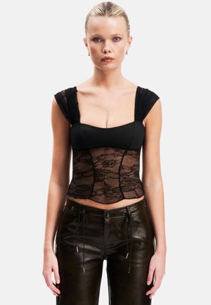 Femme aux cheveux blonds plaqués en arrière, portant un haut sans manches en dentelle et tissu noir et un pantalon en cuir noir, debout devant un fond uni.