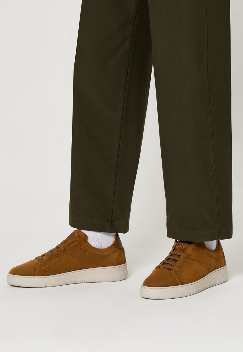 Sneaker in suede marrone con suola in gomma bianca a profilo basso, dotati di lacci rotondi e dettagli di cucitura discreti, abbinati a pantaloni verde oliva.