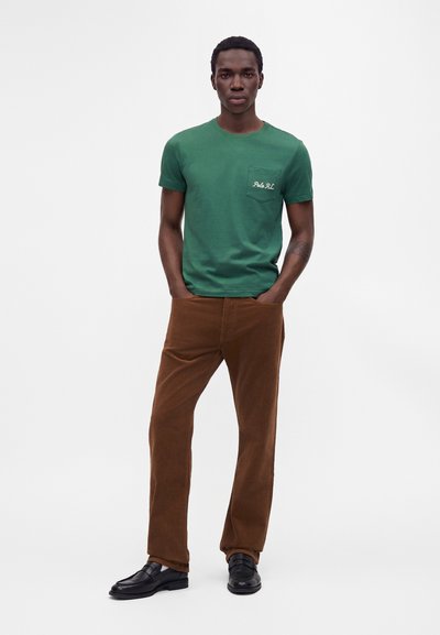 Uomo in piedi con le mani nelle tasche, che indossa una t-shirt verde con testo sulla tasca e pantaloni marroni con scarpe nere su sfondo bianco.