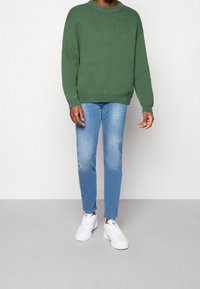 Zelený pletený svetr s kulatým výstřihem, doplněný o světle modré skinny džíny a bílé tenisky s jemnými akcentními barvami.