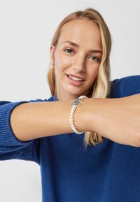 Pulsera de perlas de agua dulce con cuentas blancas y un cierre de plata, llevada en una muñeca, sobre un fondo de suéter azul.