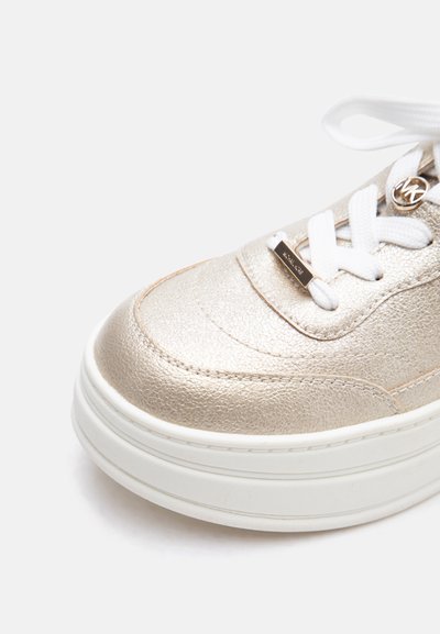 Κοντινό πλάνο του μπροστινού μέρους ενός χρυσού μεταλλικού αθλητικού παπουτσιού με λευκά κορδόνια και χοντρή λευκή σόλα πάνω σε ανοιχτό φόντο.