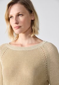 Lys beige strikket genser med vid utringning, med åpen vev tekstur og ribbede detaljer ved kragen og skuldersømmene.