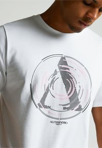 Valkoinen puuvillapaita, jossa on pyöreä graafinen kuvio mustilla ja vaaleanoransseilla viivoilla, ja brändinimi "Alessandro Zavetti" painettuna alapuolelle.