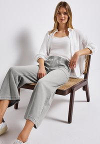 Pantalons larges gris clair avec une taille élastique, portés avec un débardeur blanc et une chemise blanche déboutonnée, assis sur une chaise en bois.