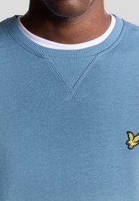 Felpa azzurra chiara con collo a girocollo a coste e un piccolo logo ricamato giallo. Texture liscia con dettagli di cucitura minimi.