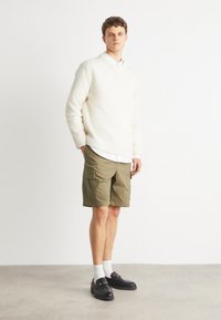 Pulover alb tricotat, pantaloni scurți cargo khaki, cămașă albă cu guler și mocasini negri cu accente aurii. Modelul stă pe un podea gri.