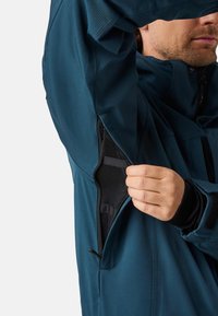 Mand, der lynlås underarmsventil med sort meshforing på blå jakke, der viser detaljer om åndbarhedsegenskaben.