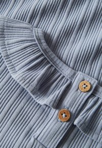 Jasnoniebieska dzianina prążkowana z falbaną i drewnianymi guzikami. Tekstura jest miękka, z widocznymi pionowymi liniami i subtelnym połyskiem.