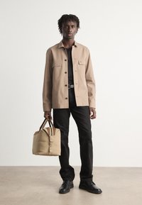 Khaki skjortejakke med to brystlommer, sort t-shirt, sorte bukser og sko. Holder en beige taske med brune læderdetaljer.