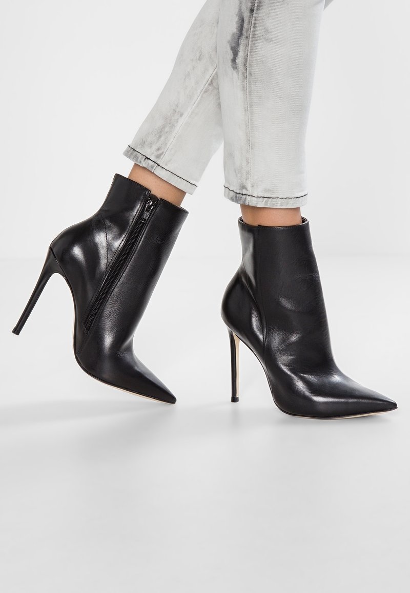 Bottes à cheville en cuir noir à bout pointu avec des talons aiguilles hauts, portées avec un jean gris clair retroussé, montrant un détail de fermeture éclair latérale.