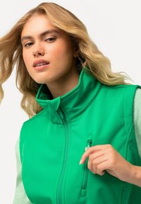 Grüne zipfeljacke mit hohem Kragen und einer kleinen Tasche. Der Stoff wirkt glatt mit matter Oberfläche. Das Modell hat lange, wellige Haare.