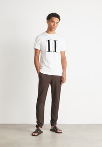 Camiseta blanca con gráfico negro "II", pantalones marrones ajustados y sandalias negras. Tela de algodón suave, diseño casual, estilo minimalista.