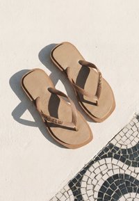 Paire de tongs beige projetant des ombres sur une surface blanche à côté d'un sol en mosaïque noir et blanc avec un motif circulaire.
