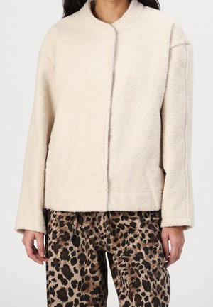 Kvinde iført en cremefarvet, fluffy jakke og leopardmønstrede bukser, der står foran en ensfarvet baggrund med hænderne afslappet ned langs siden.