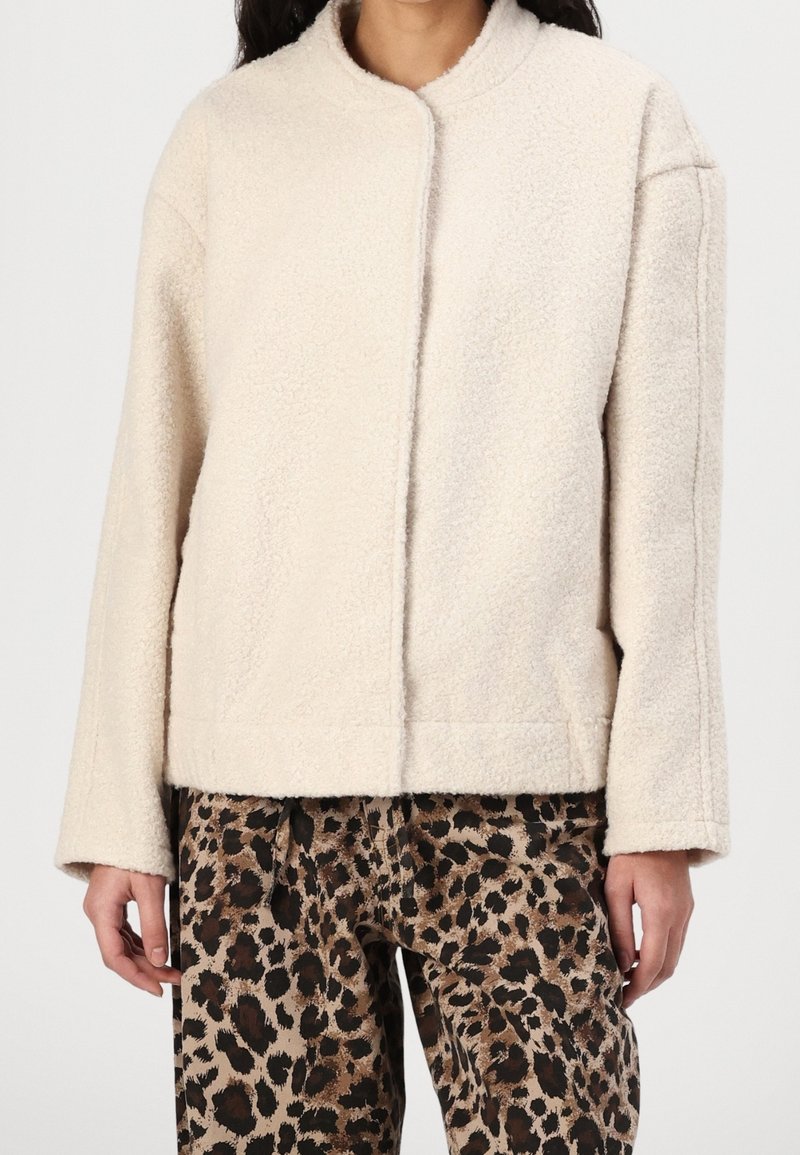 Femme portant une veste duveteuse couleur crème et un pantalon à imprimé léopard, debout devant un fond uni, les mains relâchées sur les côtés.
