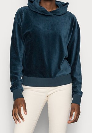 Personne portant un sweat à capuche en velours bleu foncé et un pantalon blanc, debout devant un fond uni clair.