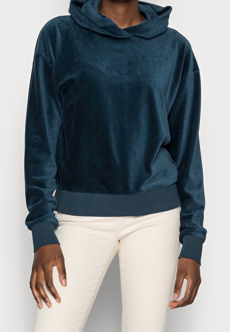 Personne portant un sweat à capuche en velours bleu foncé et un pantalon blanc, debout devant un fond uni clair.